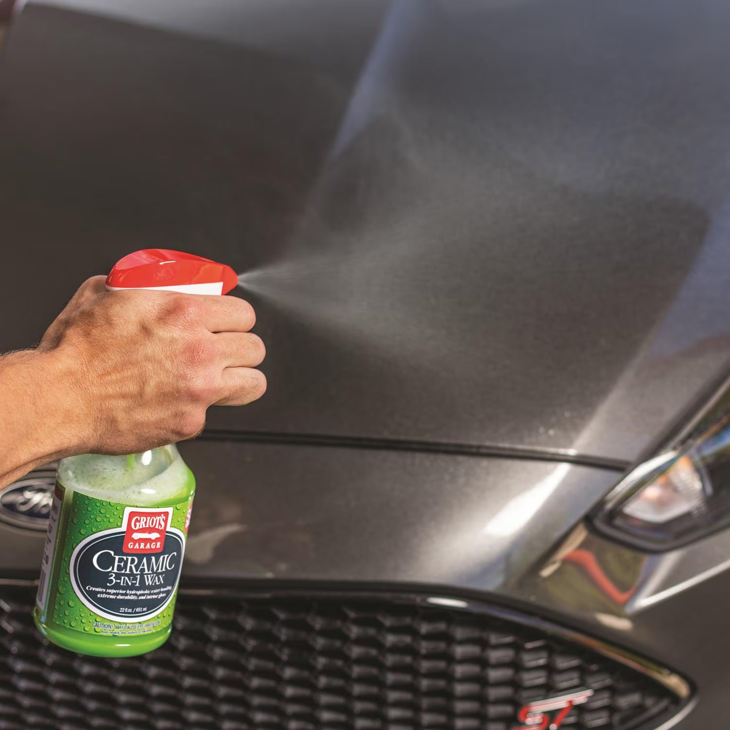 A hand sprays Griots Ceramic Shine Wash onto a dark metallic car hood, with the green and red spray bottle visible in the foreground.